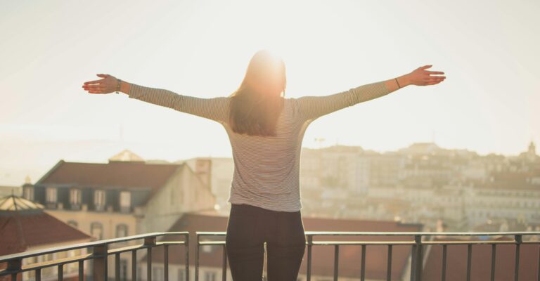 Blog 9 A woman stands with outstretched arms on a sunny balcony, embracing the morning light.