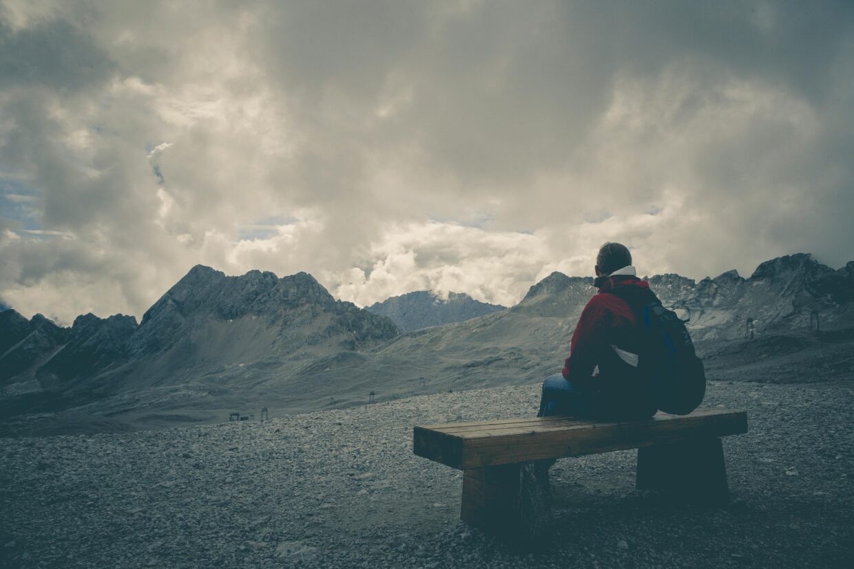 Why Do I Feel So Disconnected From Everyone? 2 A solitary traveler sits on a bench, gazing at a mountain range under a cloudy sky.