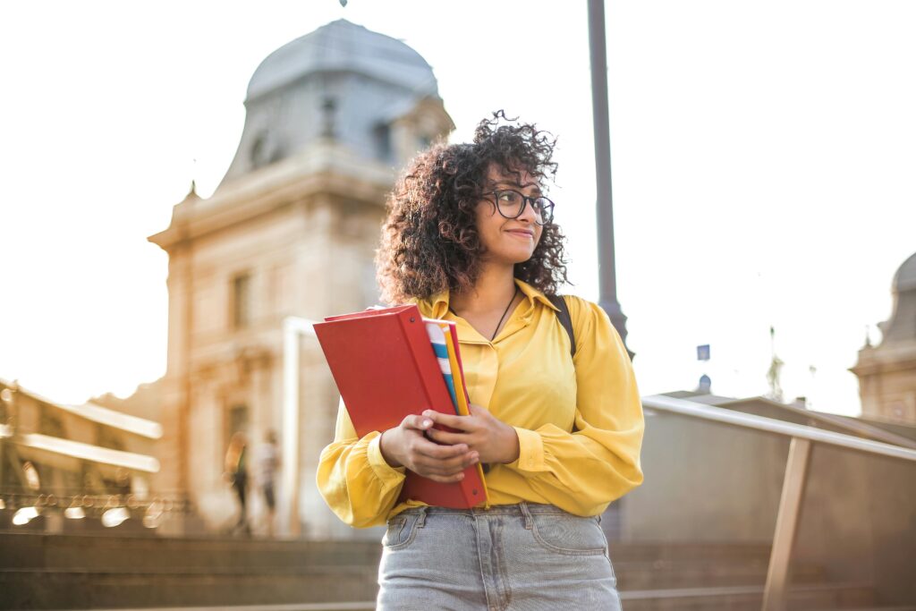 Feeling Like You Don’t Belong? Overcoming Imposter Syndrome as a Transfer Student 2 Smiling female student with curly hair and glasses holding books on campus steps.